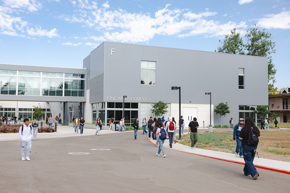 Exterior of the Fine Arts Complex at Allan Hancock College. Students walk along pathways and gather outside the modern gray building, which features large windows and a glass walkway connection, on a busy campus day under a partly cloudy sky.